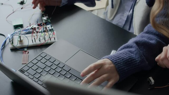 Two unrecognizable female students working on an electronics project, connecting wires to circuit board and programming it via laptop
