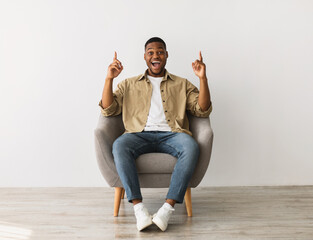 Excited African American Millennial Man Pointing Fingers Up Sitting In Chair Over Gray Wall...