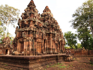 The image depicts a magnificent ancient temple complex, likely Banteay Srei, Cambodia. The central structure is a towering, multi-tiered temple with intricate carvings adorning its exterior. 