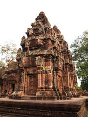 The image depicts a magnificent ancient temple complex, likely Banteay Srei, Cambodia. The central structure is a towering, multi-tiered temple with intricate carvings adorning its exterior. 