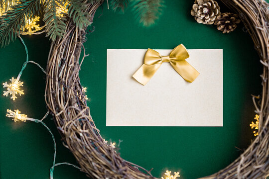Blank empty Christmas card with a yellow gold ribbon bow on a natural wooden grapevine wreath frame for Christmas decorations with soft snowflake string lights, pine cones, green fabric background DIY - Powered by Adobe