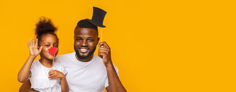 In this cheerful moment, black father and his young daughter are playfully posing together, holding fun props. The little girl waves while sporting a red nose, and father smiles broadly, copy space