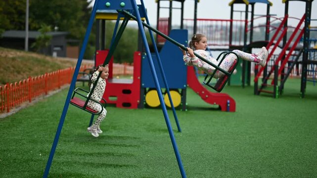 Two young sisters enjoy a beautiful day playing outside together at the park. They are happily swinging on a swing set at a colorful playground.