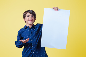 Smiling boy holds a blank billboard with one hand and points to it with the other hand. Isolated on yellow background.