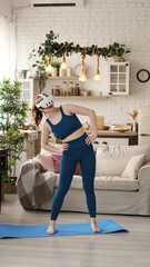 A woman in a teal outfit practices a balancing pose while wearing a VR headset in her stylish living room. This image highlights the innovative use of virtual reality technology in home fitness