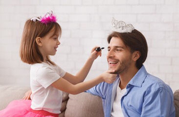 Cute little princess putting mascara on her father's eyelashes, single father enjoying time with his daughter at home