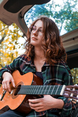 Teen girl playing guitar. Girl playing guitar in car. Autumn atmosphere. Car ride. Checkered shirt. Sun rays. Forest. Forest background. Car in forest