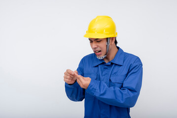 An asian worker in a yellow hard hat and blue coveralls grimaces in pain, examining a cut on his finger. Workplace accidents and safety hazards.