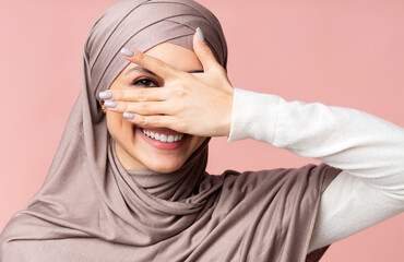 Playful arabic woman in hijab peeping through fingers and looking at camera, posing over pink background, closeup