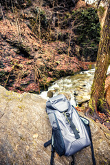 Fototapeta premium A sturdy backpack leans against a large rock near a gently flowing creek, surrounded by trees and fallen leaves, capturing the essence of a peaceful outdoor escape