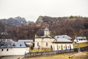 Beneath a gray sky, a tranquil monastery stands amid rolling hills and rugged mountains, adorned...