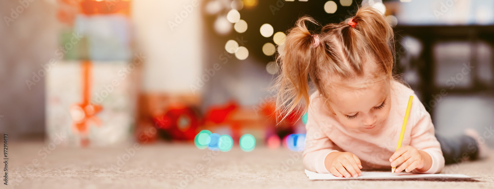 Wall mural Cherished wish. Little girl drawing letter to Santa Claus, lying on floor near Christmas tree, empty space