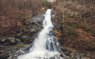 Obraz premium Rushing water tumbles gracefully over rugged stones, while surrounding trees display vibrant autumn colors, creating a serene natural escape away from urban life