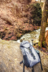 An inviting hiking area unfolds by a bubbling river, with a gray backpack settled on a smooth rock amidst the rustling leaves and towering trees
