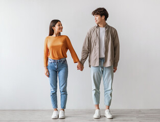 Full length portrait of millennial Asian couple smiling and looking at each other, holding hands, standing against white studio wall. Affectionate young man and woman being in love