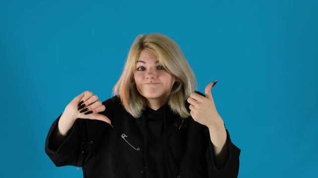 Young woman hesitantly raises one thumb up and other down. Lady struggles to decide with mixed signals reflecting indecision on blue