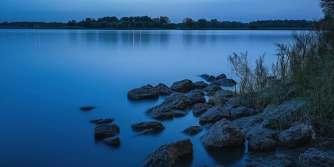 A tranquil lakeside scene during blue hour, with rocks lining the calm water and soft light reflecting across the surface.