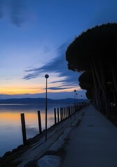 A quiet lakeside path lined with trees and lampposts, bathed in the fading light of dusk as the sky reflects over the calm waters below.