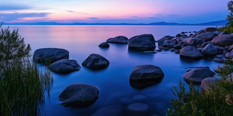 A stunning twilight sky reflects vibrant colors over the calm water, with large rocks scattered along the peaceful shoreline.
