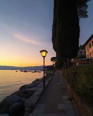 A tranquil lakeside walkway illuminated by warm lampposts, bordered by trees and rocks, as the sun sets, casting a soft glow over the water.