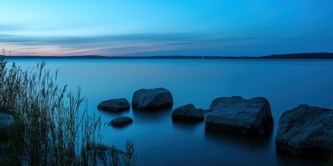 Massive rocks rest in still waters at dusk, with tall reeds along the shoreline and the horizon lit by soft, fading light.