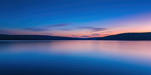A serene sunset casts soft colors over a still lake, with smooth reflections on the water and distant hills in the background.