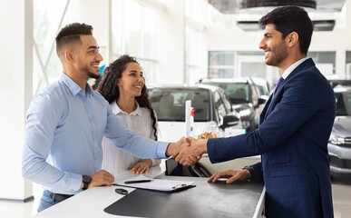 Smiling middle-eastern young man and woman buying car at auto salon, handsome arab guy shaking sales assistant hand, making successful deal, happy family got brand new luxury car, side view