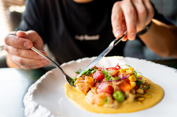 person dining, focusing on hands using utensils to eat colorful gourmet dish with vibrant garnishes on a white plate. setting is elegant, emphasizing culinary presentation and fine dining experience