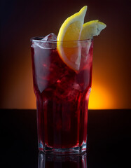 Сocktail with cherry juice and lemon slices on a black surface, against a dark sunset background