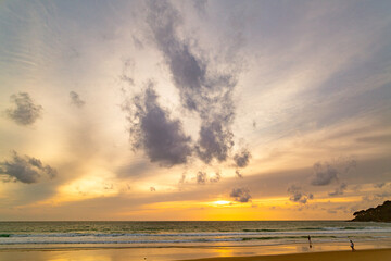 A stunning sunset over the ocean with soft golden light reflecting on the water, featuring silhouettes of people walking along the shore. Water flows from a small stream into the sea.