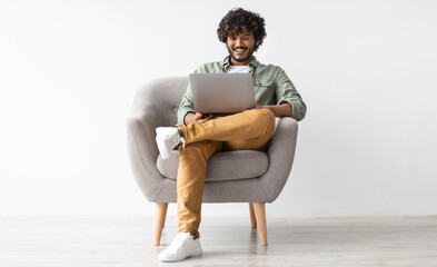 Joyful young indian guy in casual sitting in armchair, chilling with modern laptop, gambling or...