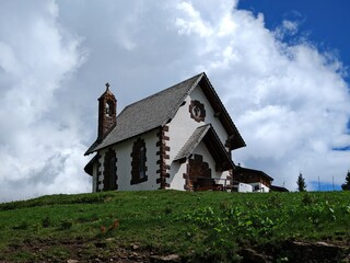 Fototapeta premium White Chapel on a Green Hilltop, Passo Rolle - A stunning view of a charming chapel nestled amidst the breathtaking Dolomites