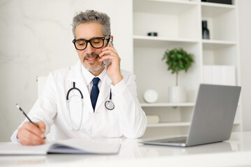 A senior male doctor with grey hair is talking on the phone while writing notes. The setting...