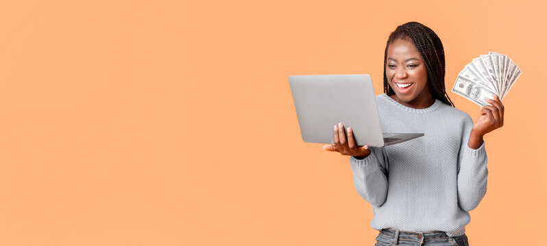 A joyful young African American woman is smiling broadly as she holds a stack of cash in one hand and a laptop in the other. She appears to be celebrating a financial achievement, copy space