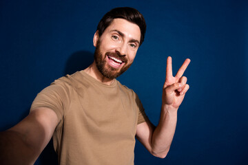 Selfie of young handsome blogger guy brunet hair model in beige t shirt showing v sign into cadre...