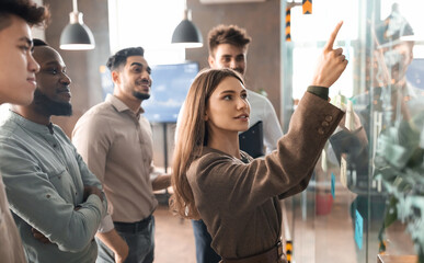 Portrait of young businesswoman mentor coach leader writing idea or task pointing at sticky notes on glass wall, diverse team developing work plan in creative corporate office at stand up meeting