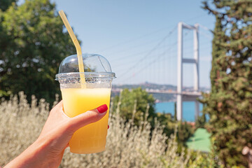 A refreshing glass of orange lemonade in hand against the backdrop of bosphorus bridge, capturing the essence of summer in Istanbul