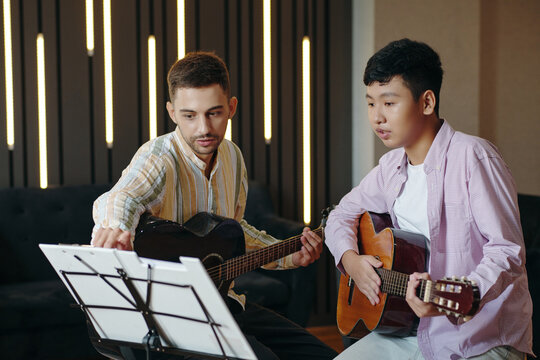 Young men playing guitar and reading sheet music in modern studio with vertical lights on wall background. Focusing on learning guitar technique and musical collaboration