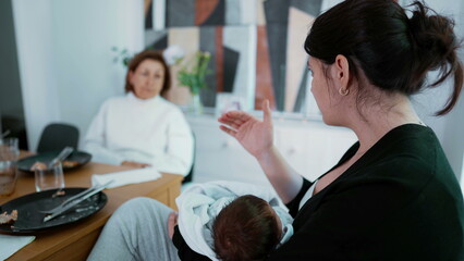 Mother holding baby while having an animated discussion with an elderly woman at a dining table, highlighting family dynamics and generational bonding in a cozy home environment