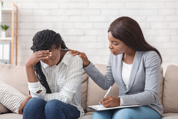 Female psychologist consoling depressed black woman at therapy session in office, supporting her to...