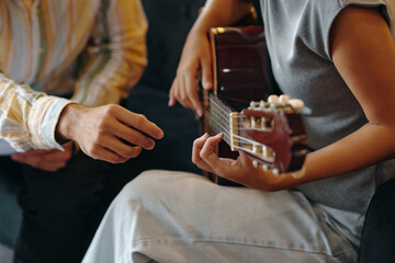 Hands of person strumming guitar strings during music lesson, another person assisting by adjusting...