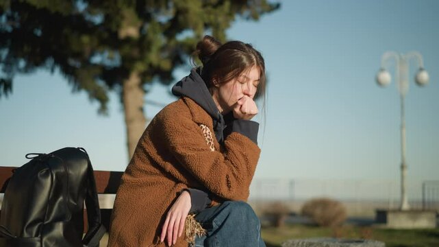 A girl sits alone on a bench in an urban park, overwhelmed with grief, her hand resting on her face. She wears a brown coat and blue jeans, with a black backpack placed beside her