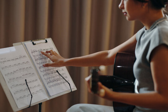 Person playing acoustic guitar while reading sheet music placed on stand Musician actively engaged in practicing or performing with curtains in the background