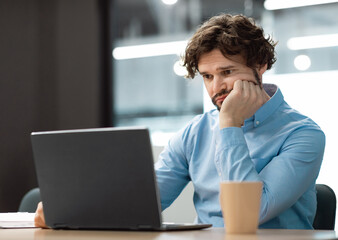 Portrait of sad bored business man sitting at desk using pc, leaning head on hand looking at...
