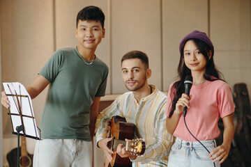 Three friends practicing music in studio setting, featuring an Asian teen holding sheet music, a Caucasian man playing guitar, and a young girl singing into microphone © DragonImages