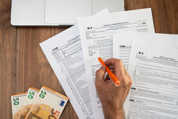 Obraz premium Close-up of a person filling out tax forms on a wooden desk with an orange pen. Euro bills and a laptop are nearby, representing financial calculations and tax preparation. tax season, accounting