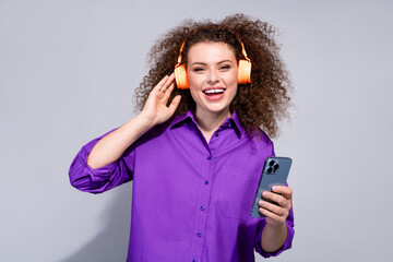 Portrait of pretty young woman touch headphones hold phone wear violet shirt isolated on grey color background