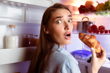 Cheat meal concept. Scared young woman eating croissant near fridge late in night
