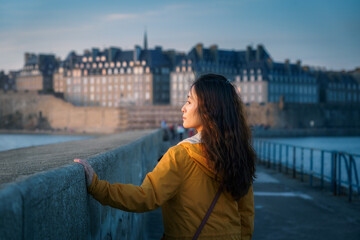 An Asian woman walking on the Saint Malo bords de La Rance, against Saint-Malo historic city center at dusk