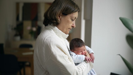 Obraz premium Elderly woman gently cradling a newborn baby, standing by a window in a softly lit room, embodying tenderness and care as she looks down at the infant with a calm and nurturing expression